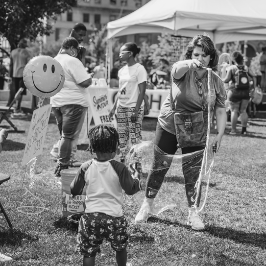 black and white image of an outing with a kid and a woman blowing a huge bubble outdoors